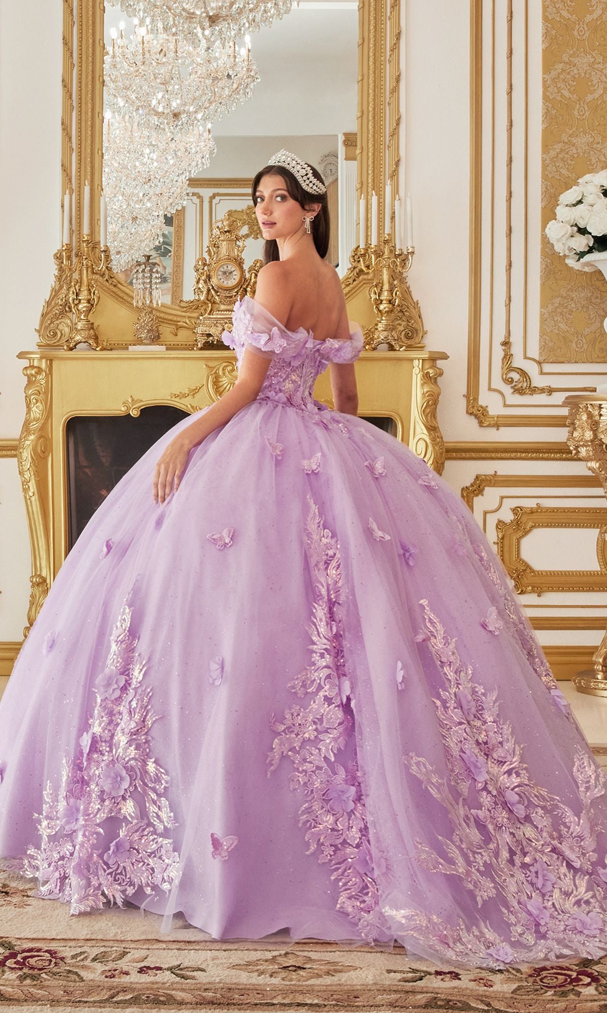 A woman in the Quinceanera Dress 15713 by Ladivine, a lavender off-the-shoulder gown with floral and butterfly applique, stands in an ornate gold-trimmed room wearing a tiara and looking over her shoulder.
